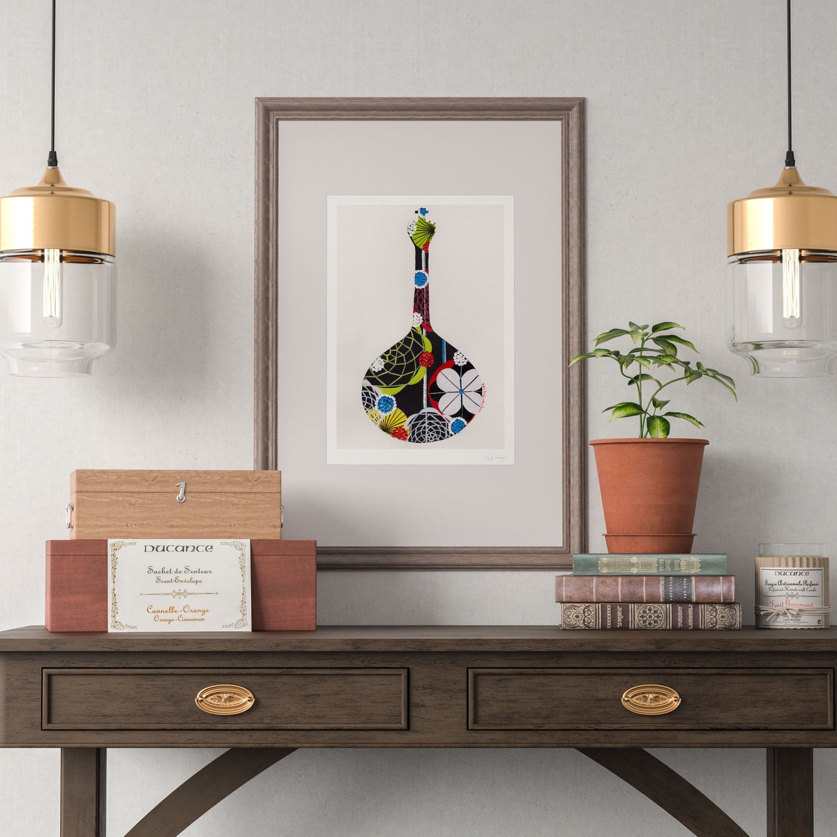 A framed fine art print, by Lis Gonçalves. Open edition, signed by the artist (printed in Hahnemühle matt fibre paper) featuring a colorful Portuguese guitar is displayed on a wooden console table in a styled room setting with hanging pendant lights, a potted plant, and books.