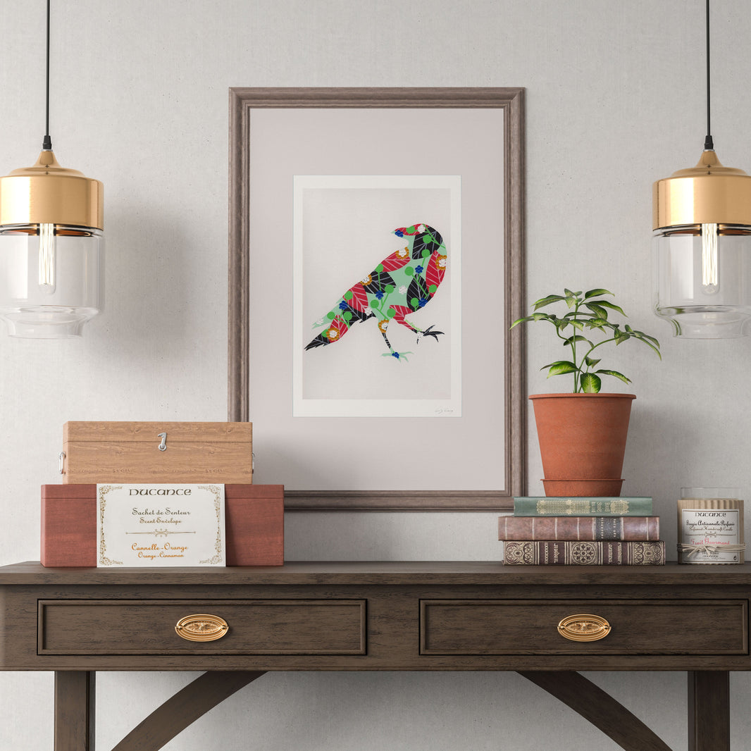 A framed fine art print, by Lis Gonçalves. Open edition, signed by the artist (printed in Hahnemühle matt fibre paper) featuring a colorful watercolor-style raven on a light background, displayed on a dark wood console table in a styled room setting with hanging pendant lights, books, and a potted plant.