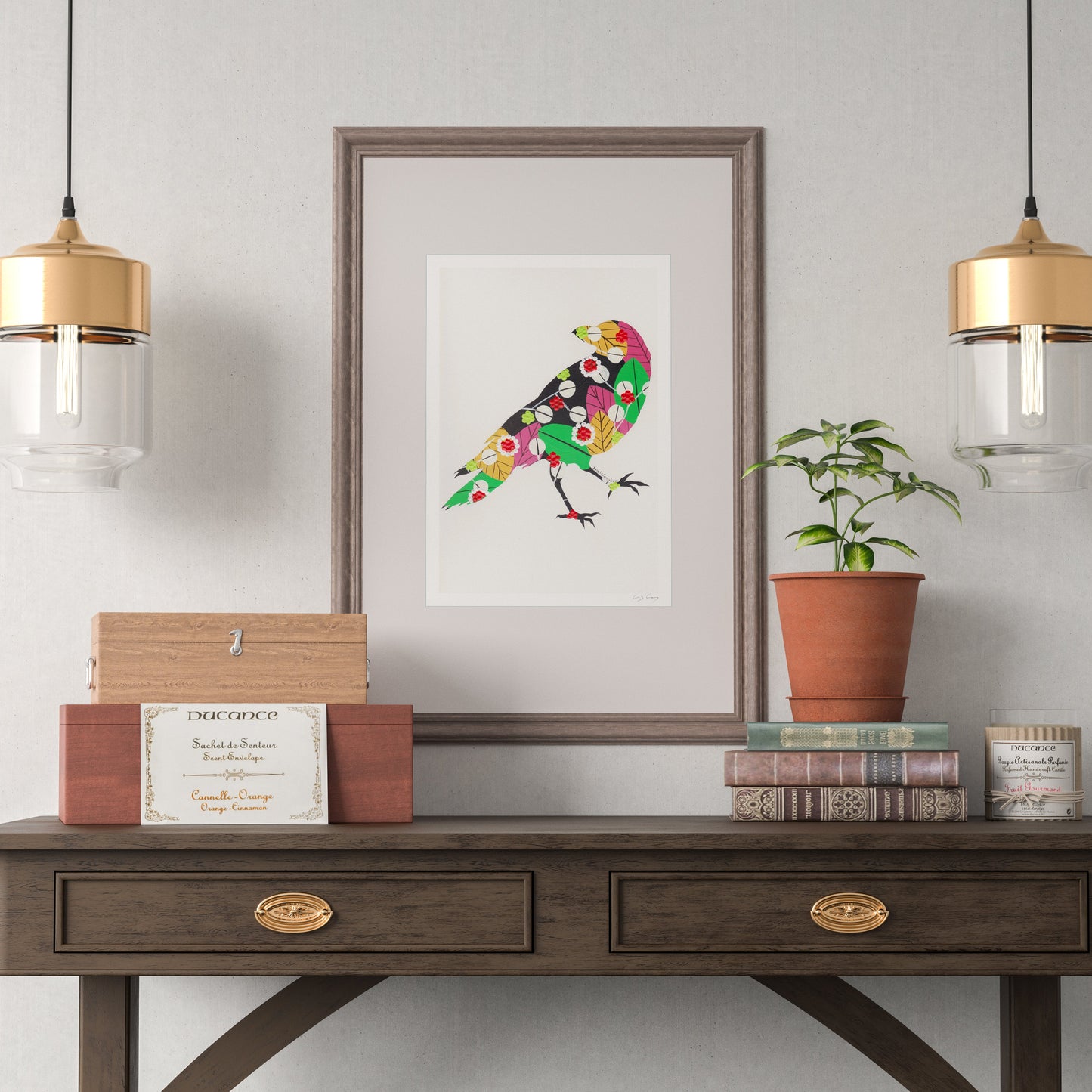 A framed fine art print, by Lis Gonçalves. Open edition, signed by the artist (printed in Hahnemühle matt fibre paper) featuring a colorful raven hanging on a light gray wall above a dark wood console table. The scene is styled with two gold and clear glass pendant lights, a small potted plant, books, and decorative boxes.