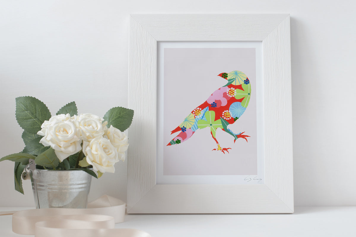 A framed fine art print, by Lis Gonçalves. Open edition, signed by the artist (printed in Hahnemühle matt fibre paper) featuring a colorful raven on a white surface with flowers and candles.