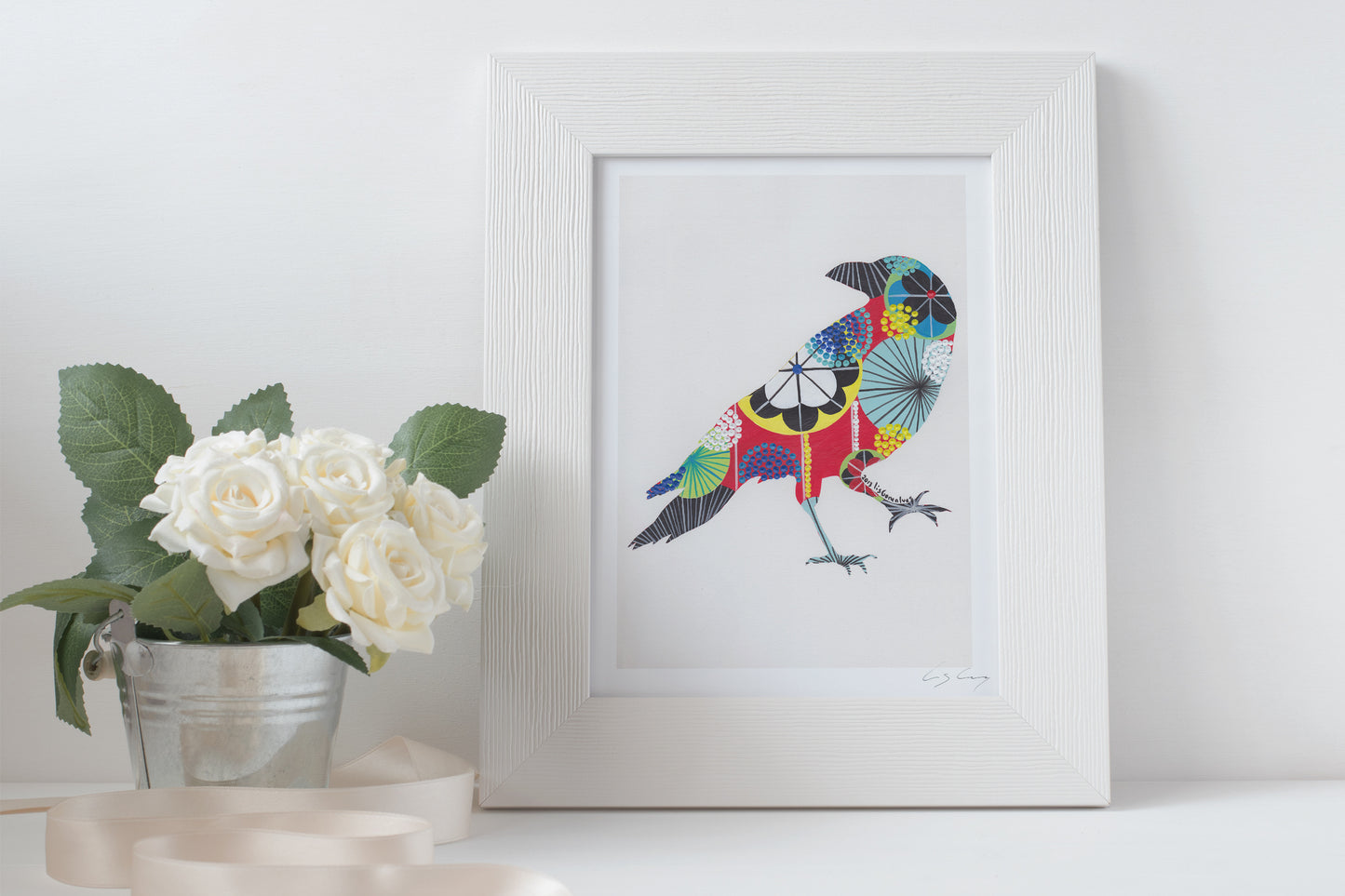 A framed fine art print, by Lis Gonçalves. Open edition, signed by the artist (printed in Hahnemühle matt fibre paper) of a colorful raven illustration on a white surface with flowers and candles.